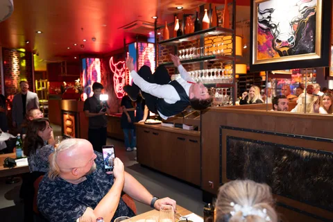 Ein Showkellner zeigt einen Salto neben den Restauranttischen, während Gäste zuschauen und filmen.