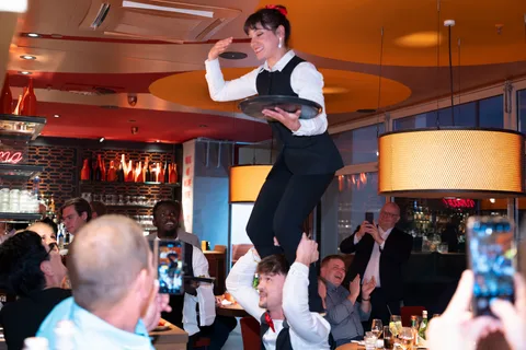 Eine Showkellnerin mit Tablett auf den Schultern ihres Kollegen, umringt von Gästen.